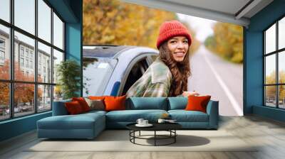 Beautiful woman takes pictures on the camerafrom the car window. Smiling woman enjoying autumn weather. Rest, relaxation, lifestyle concept. Wall mural