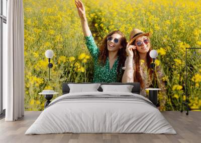 Cheerful girlfriends are walking in a blooming rapeseed field in sunny weather. Two young women enjoying and having fun outdoors. Nature and fun concept. Wall mural