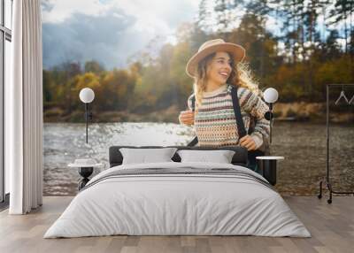 Female traveler in hiking boots with a backpack and a hat explores nature. A woman stands on the beach near the river enjoying the scenery and sunny weather, feeling freedom. Wall mural