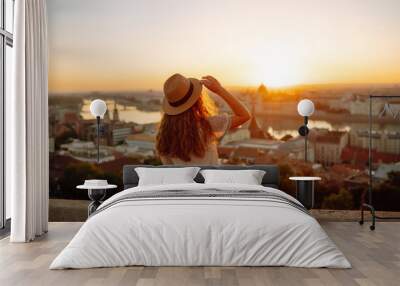 Happy traveler in a stylish dress and hat enjoys the sunrise or sunset with stunning views of the city. Back view. Lifestyle, travel, tourism, nature, active lifestyle. Wall mural