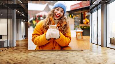 Happy woman drinks a hot drink at a Christmas market decorated with festive lights. Festive Christmas fair, winter holidays concept. Wall mural