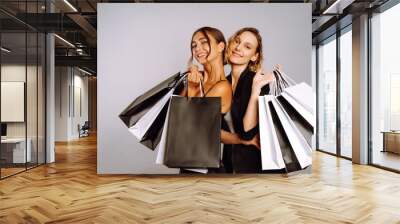 Two fashion women in black dresses with shopping bags. Young women friends with black and white bags posing on grey background. Purchases, black friday, discounts, sale concept. Wall mural