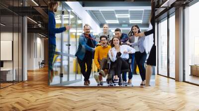 Young colleagues group having fun together, riding on chairs in office, diverse excited office workers enjoying break, laughing, engaged funny activity, celebrating corporate success. Wall mural