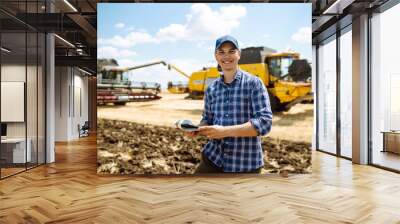 Young farmer with tablet standing in front of combine harvester  in field. Agriculture, gardening or ecology concept. Wall mural