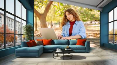 Young  freelancer woman in casual attire focused on laptop while sitting at a park table on a sunny day, enjoying a moment of productivity. E-learning concept. Remote work. Wall mural