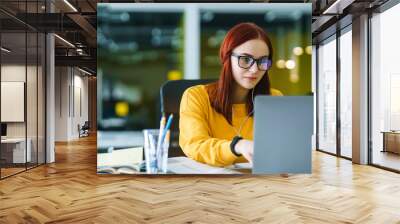 Young girl works at a computer in the office. Beautiful freelancer working from a laptop computer. Teenager student at the computer listens music to earphones and prepares for exams. Business woman. Wall mural