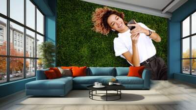 Young woman sitting on the grass  in a city park and using a smartphone. Technology and modern lifestyle concept. Wall mural