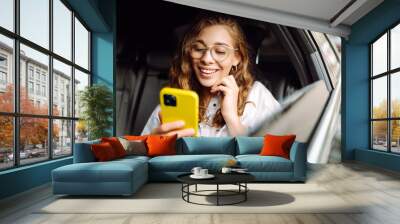 Young woman uses a smartphone while sitting in the back seat of a car. Female happy in car traveling on the road to destination. Technology, blogging, business concept. Wall mural