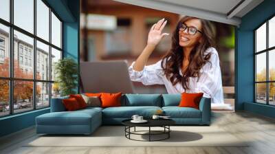 Young woman with wireless headphones calling on laptop, talk by webcam, video conference sitting on modern cafe. Concept for education, business, blog or freelance. Wall mural