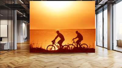 silhouette of 2 two person - man and woman. Side view of couple riding on seashore with their bicycles. sunset sky on background. couple fallen in love. Wall mural