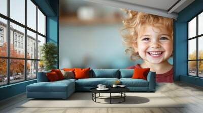 A cheerful toddler with curly hair beams with a wide smile while wearing a red top, standing playfully indoors with soft background lighting, radiating pure happiness. Wall mural