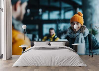 A cheerful woman wearing an orange beanie and winter attire smiles brightly at a café counter, as she enjoys a warm beverage in a cozy, modern coffee shop environment. Wall mural