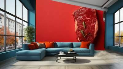 A fresh, uncooked steak sits on a bold red background with a fork nearby, seasoned with sprigs of rosemary and pepper, highlighting its culinary appeal. Wall mural