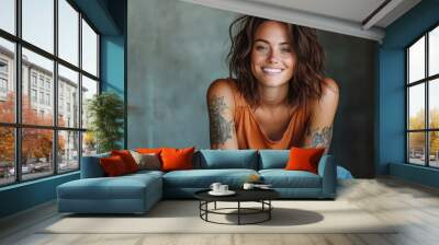 A happy woman adorned with tattoos grins widely in a casual orange top, set against a textured background, reflecting a carefree and modern spirit. Wall mural