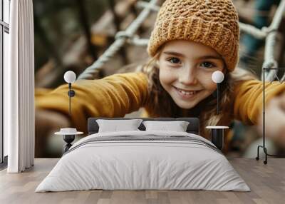 A happy young girl wearing a knitted hat and yellow sweater enjoys climbing a rope structure in an outdoor setting, captured with a close-up of her smiling face. Wall mural