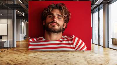 A man with tousled hair lies against a vibrant red wall, wearing a striped shirt, presenting a relaxed and confident demeanor, blending casual and bold styles. Wall mural