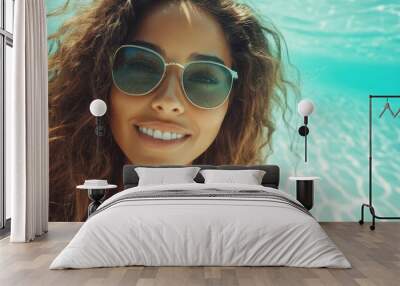 A smiling woman, with curly hair and sunglasses, enjoys a refreshing dip underwater, capturing a carefree and sunny vacation moment against clear blue water. Wall mural