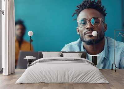 A stylish young man with glasses sits confidently in a relaxed pose wearing a light blue casual outfit, evoking a sense of ease and contemporary flair. Wall mural