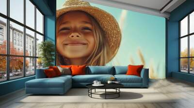 A young girl smiles warmly, captured in a field with a clear blue sky, wearing a straw hat. Her happiness is a perfect reflection of natural beauty and innocence. Wall mural