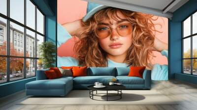 A young girl with curly hair, wearing a blue sweatshirt, sunglasses, and a cap, poses against a pink background, expressing modern casual style and attitude. Wall mural