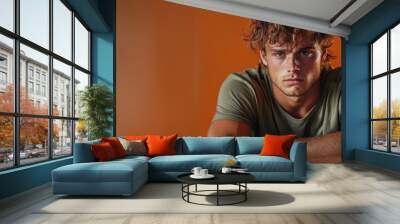 A young man with messy hair sits pensively against a vibrant orange background, wearing a casual t-shirt, exuding a sense of introspection and youthfulness. Wall mural