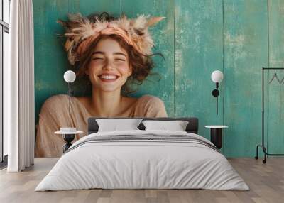 A young woman with a radiant smile, freckles, and a feathered headpiece, dressed in a natural-toned outfit, leaning against a teal wooden background. She exudes joy. Wall mural