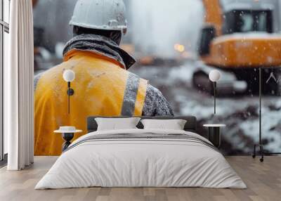 The back view of a construction worker with a white helmet and orange high-visibility jacket, standing in a snow-covered construction site, focuses on heavy machinery. Wall mural