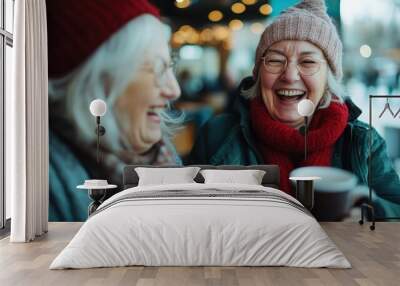 Two joyful older women share a hearty laugh over steaming cups of coffee at a cozy café setting, capturing a moment of friendship and warmth during winter. Wall mural