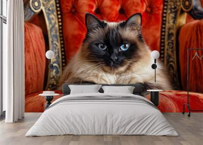 A cat is sitting on a red chair with its paws on the arm. The cat has a blue eye and is looking at the camera. The chair is a large, ornate piece of furniture with gold accents. The scene is cozy Wall mural