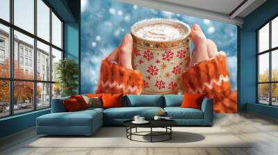 A person is holding a mug with a snowflake pattern on it. The mug is filled with hot chocolate, and the person is wearing an orange sweater. The image conveys a cozy and warm atmosphere Wall mural