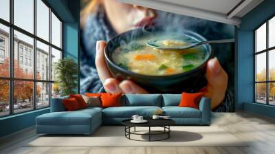 A woman is holding a bowl of soup and blowing on it. The soup is hot and steamy, and the woman is trying to cool it down. Concept of warmth and comfort Wall mural