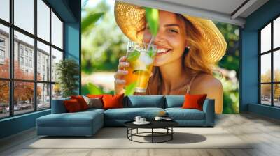 A woman is smiling and holding a glass of a drink with a straw in it. She is wearing a straw hat and is sitting in a green area Wall mural