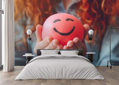 Close-up of hands holding a red stress ball with a smiley face, promoting happiness and stress relief. Wall mural