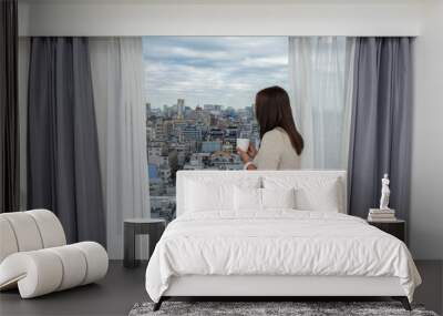 Stay home Stay safe, young woman holding a cup of coffee in the morning at home and opening window curtains, looking out window with beautiful cityscape view from the bedroom, blue sky. Wall mural