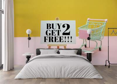 Buy 2 Get 2 Free!!! Sale promotion text on easel with trolley shopping cart on yellow and pink background Wall mural