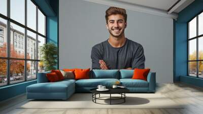 A handsome young man smiling with his arms crossed against a grey background, wearing casual with a confident and happy expression in a studio portrait. Wall mural