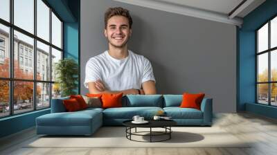 A handsome young man smiling with his arms crossed against a grey background, wearing casual with a confident and happy expression in a studio portrait. Wall mural