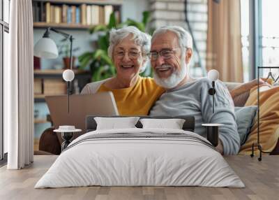 A happy senior couple sits together at a laptop, looking into the camera and smiling, websurfing on internet with laptop at home and studying the laptop screen intently Wall mural