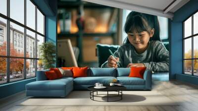 A young Asian girl is sitting at a table, smiling and writing in her notebook with a green pencil. Asian girl student sits and writes note in the classroom, Education in schools in the, kindergarten. Wall mural