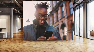 Photo of a happy man looking at his phone on the street in an urban setting. He is smiling and wearing modern clothing. The background features buildings Wall mural