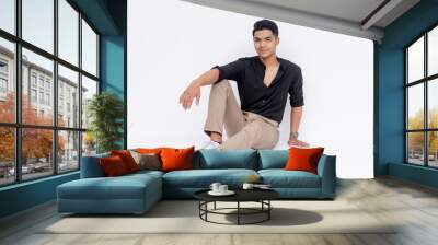 A relaxed and stylish young man sits casually with a confident smile, dressed in smart casual attire against a white background. Wall mural