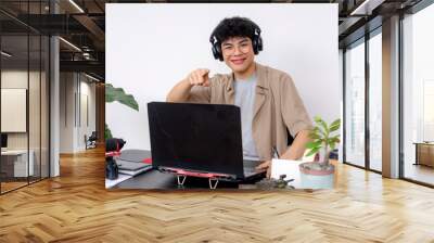 Happy young Asian graphic designer with headphones working on a laptop in a creative office space. Pointing at the camera. Wall mural