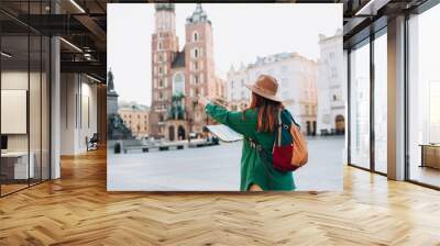 Attractive female tourist is exploring new city. Redhead woman pointing finger and holding a paper map on Market Square in Krakow. Traveling Europe in autumn. St. Marys Basilica. Active lifestyle Wall mural