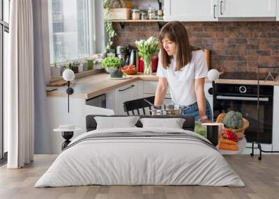 Close up woman in white t-shirt cooking soup with fresh vegetables in kitchen at home. Menu, recipe book banner. Girl reads the recipe in laptop. Caucasian model using internet in loft apartment. Wall mural