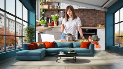 Close up woman in white t-shirt cooking soup with fresh vegetables in kitchen at home. Menu, recipe book banner. Girl reads the recipe in laptop. Caucasian model using internet in loft apartment. Wall mural