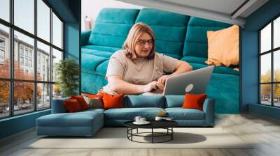 Nice beautiful woman in glasses working at the notebook near the sofa at home. Concepts of education, work and comfort. 30s female working indoors Wall mural