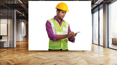 Indian male architect wearing yellow helmet and safety jacket writing something using clipboard while standing in studio Wall mural