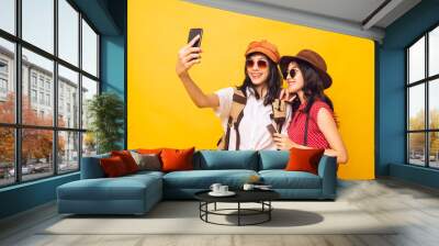 Two Asian pretty young girl smiling wear sunglasses and take a selfie by smartphone together, Young women backpacker take a selfie and holding passport in studio yellow background. Wall mural