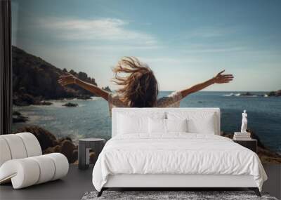 Woman with outstretched arms enjoying the wind and breathing fresh air on the rocky beach, back shot, summer time Wall mural