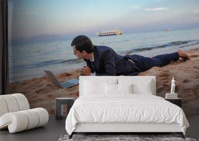 lateral view of a young man in suit with laptop lies on the beach working on the seaside Wall mural